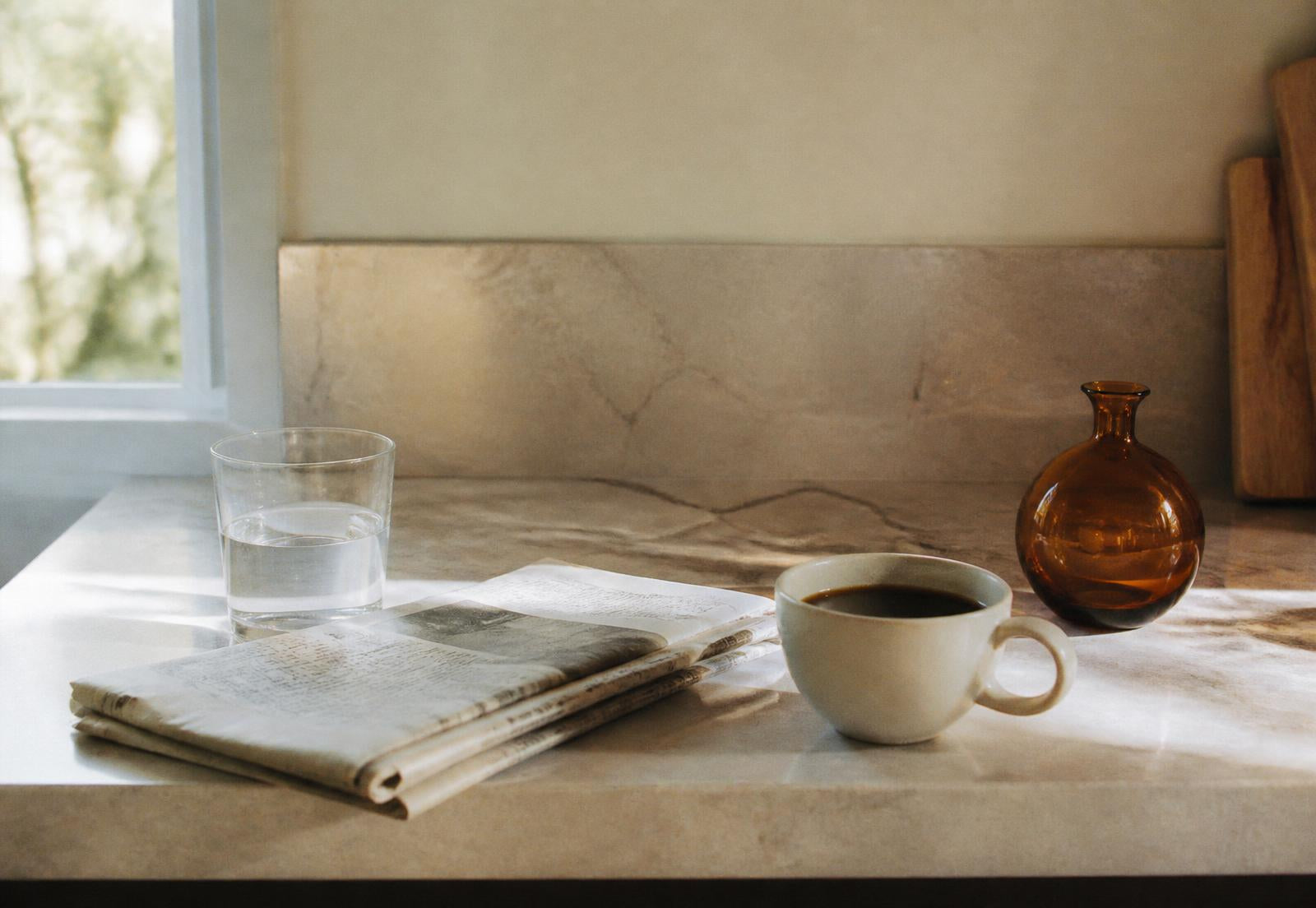 Morning light across a kitchen counter with a glass of water, folded newspaper, coffee and a small amber vessel.